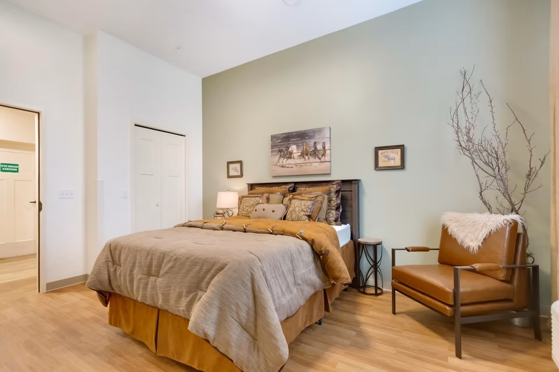 A cozy bedroom with a neatly made bed featuring multiple pillows and a beige comforter. On the wall above the bed are three framed pictures, including one of running horses. To the right of the bed is a brown leather armchair with a white fur throw draped over the back, and a tall vase with decorative branches. The room has light wood flooring and light-colored walls, with a door and closet visible on the left side.