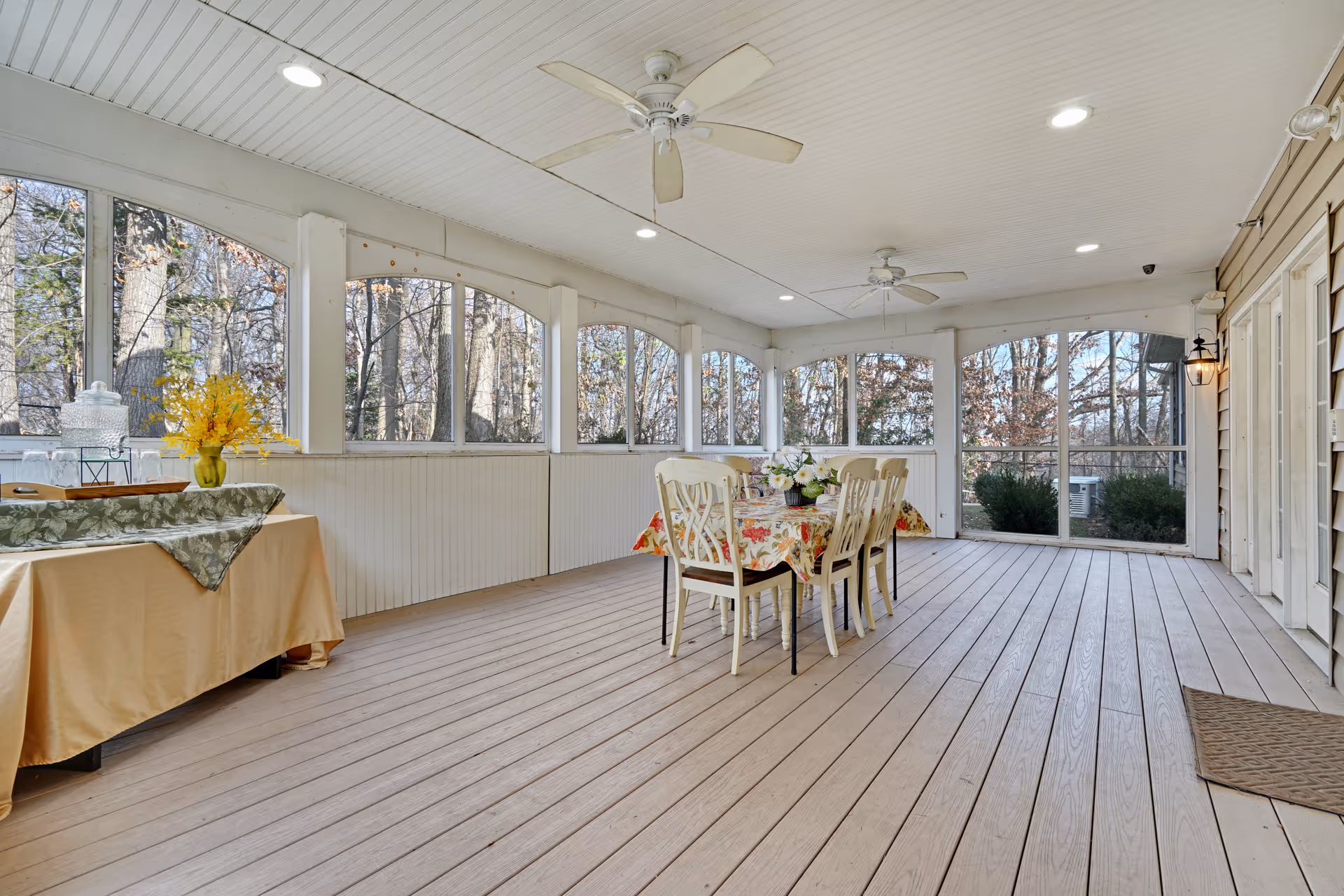A spacious screened-in porch with wooden flooring and white ceiling fans. There is a table with six chairs covered with a floral tablecloth in the center, and a side table with a yellow flower arrangement and a water dispenser on the left. The porch is surrounded by large screened windows showing a view of trees outside.