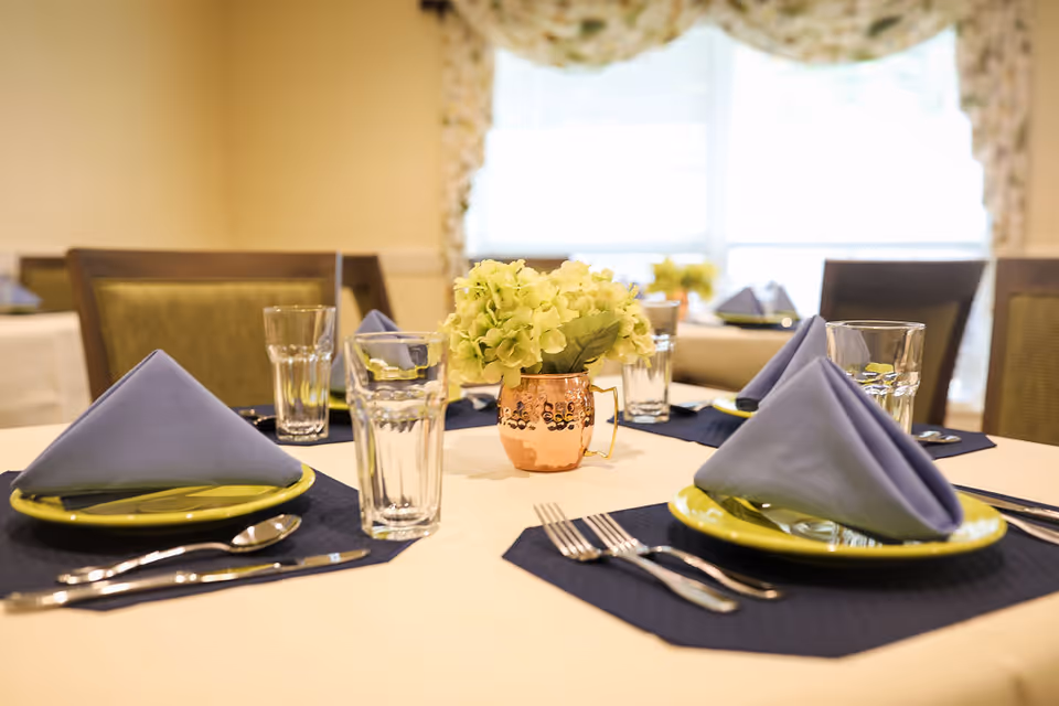 A dining table set with folded blue napkins, glasses, utensils, and a small floral centerpiece in a bright dining room.