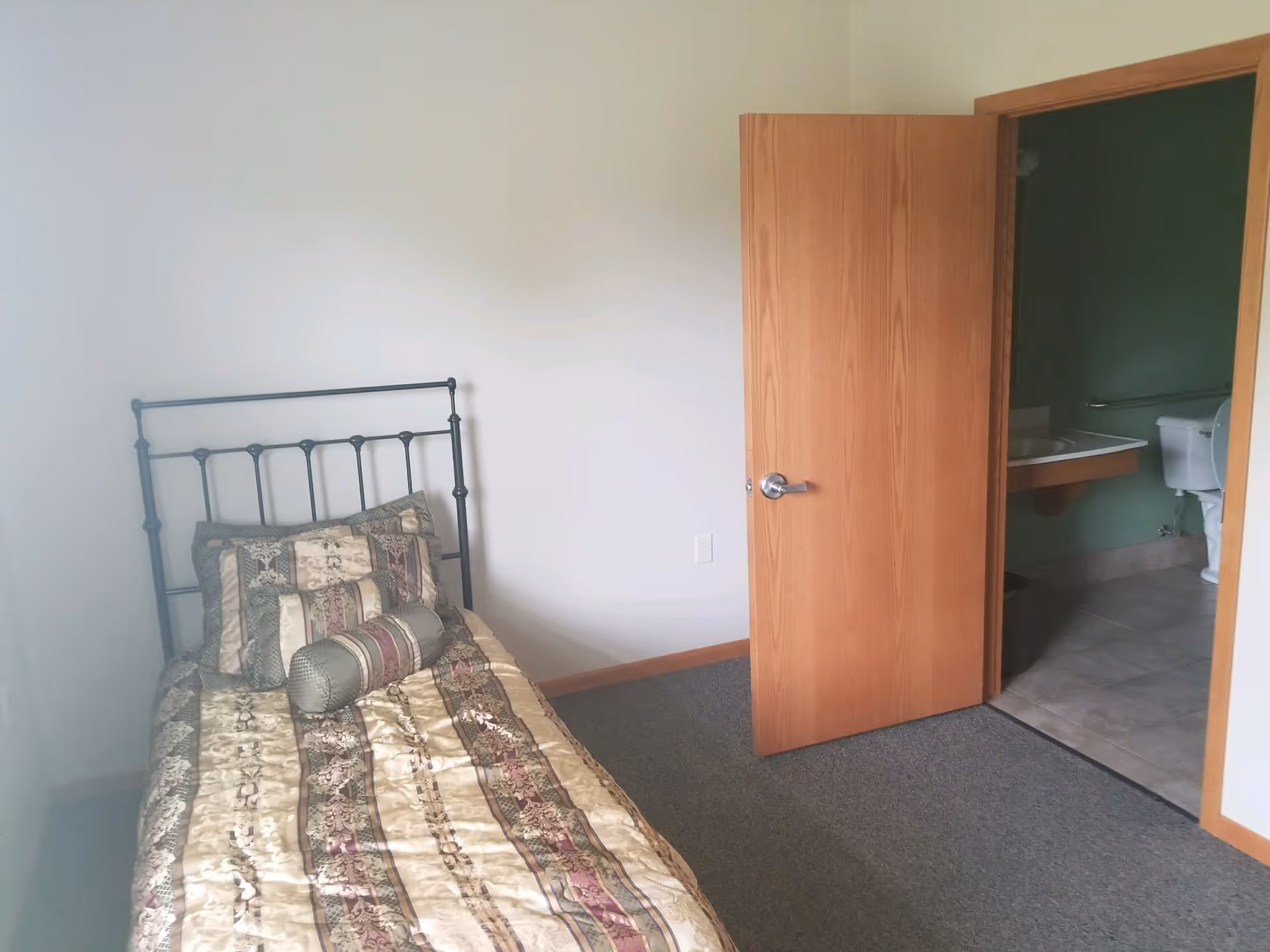 A small bedroom with a single bed featuring a decorative patterned bedspread and pillows. The room has plain white walls and a carpeted floor. An open wooden door reveals an adjacent bathroom with a toilet, sink, and tiled floor.
