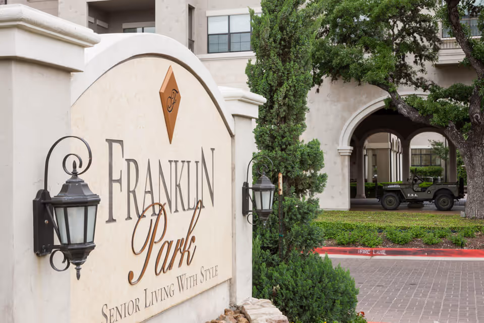 Entrance sign for Franklin Park Senior Living with decorative lamps on either side, surrounded by greenery and trees, with a building and a vintage military-style jeep visible in the background.