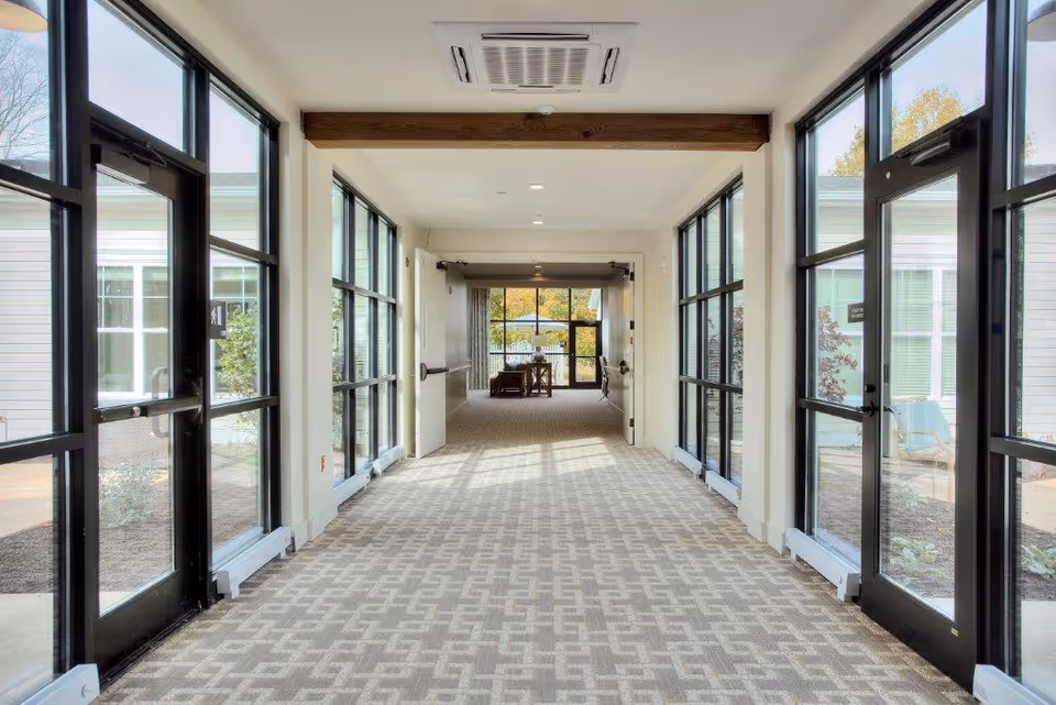 A bright hallway with large windows and glass doors on both sides, leading to a seating area with chairs and a table at the far end. The floor is carpeted with a geometric pattern, and there is a wooden beam on the ceiling along with recessed lighting.