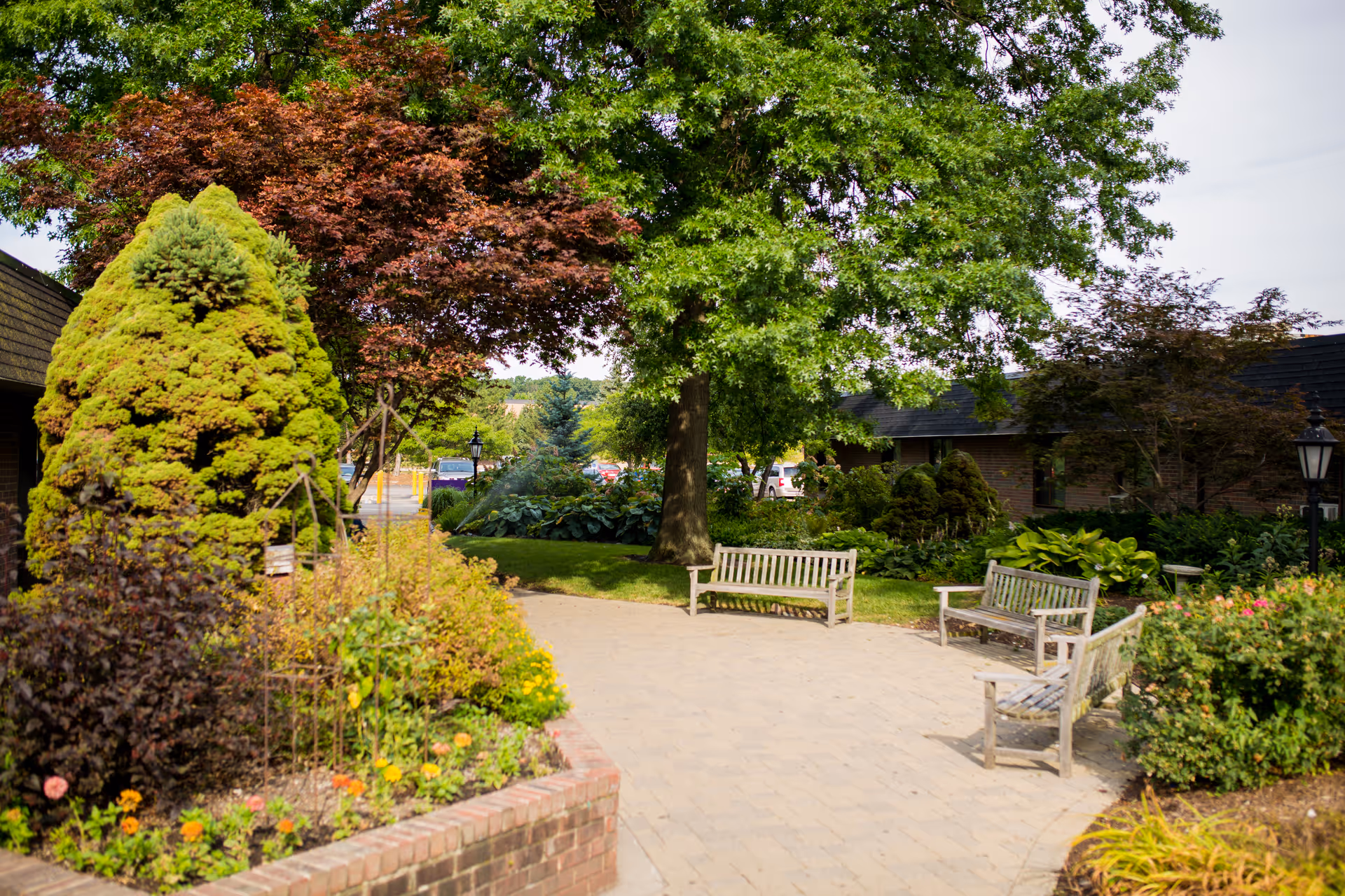 A peaceful outdoor garden area at Westland, a Villa Center, featuring a paved walkway surrounded by lush greenery, colorful flowers, and several wooden benches under large trees.