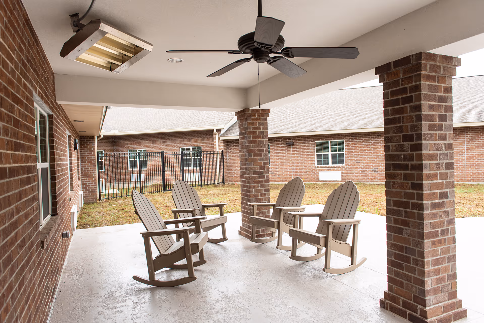 Covered outdoor patio area with four wooden rocking chairs arranged in a circle on a concrete floor. The patio is supported by brick columns and has a ceiling fan and a heater mounted on the ceiling. In the background, there is a fenced grassy courtyard surrounded by brick buildings with windows.