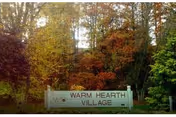 Entrance sign that reads "Warm Hearth Village" set in front of trees with autumn foliage.