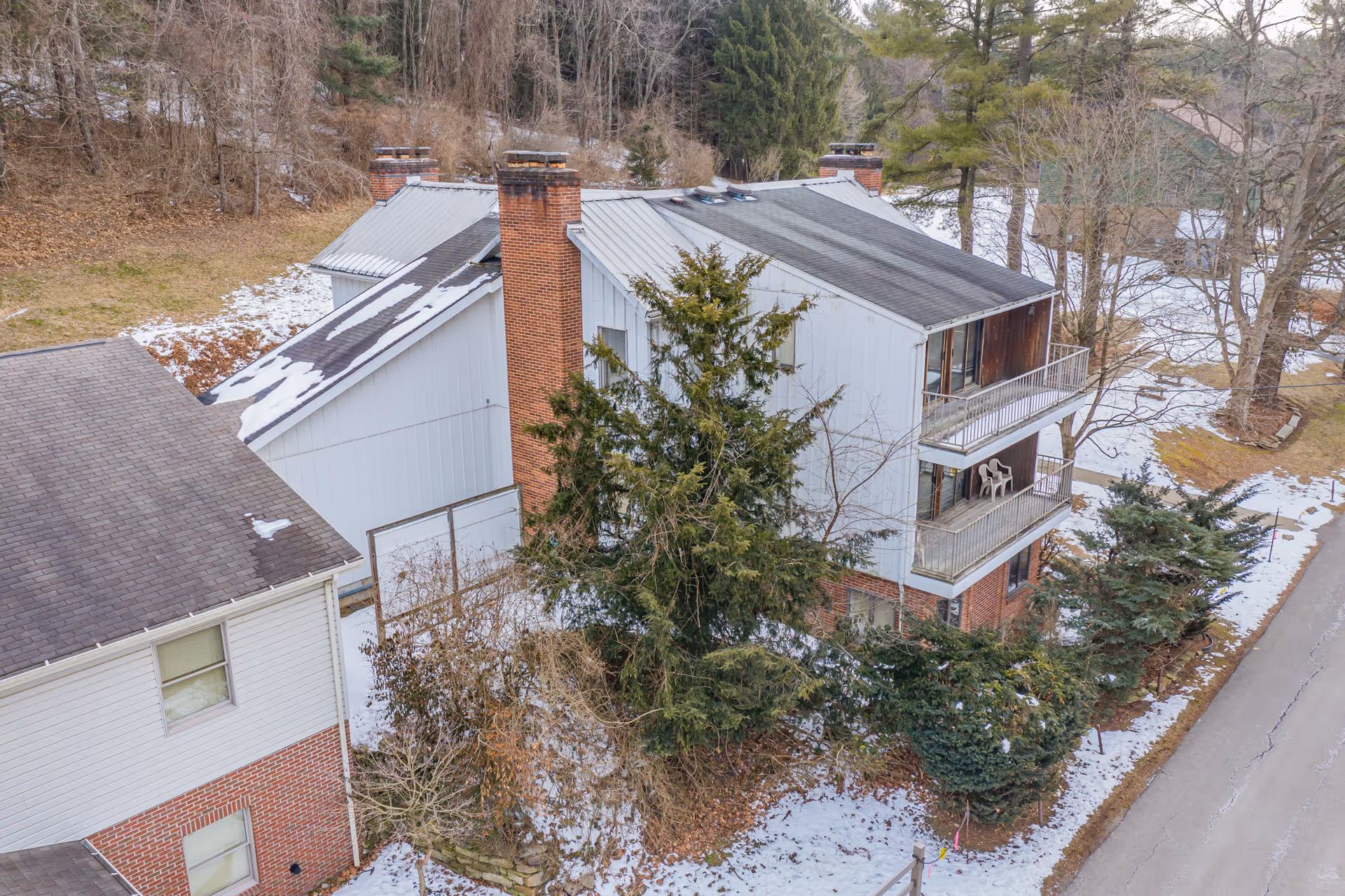 A multi-story residential building with balconies, surrounded by trees and patches of snow on the ground. The building has a mix of brick and light-colored siding with a sloped roof and multiple chimneys. A road runs alongside the building.