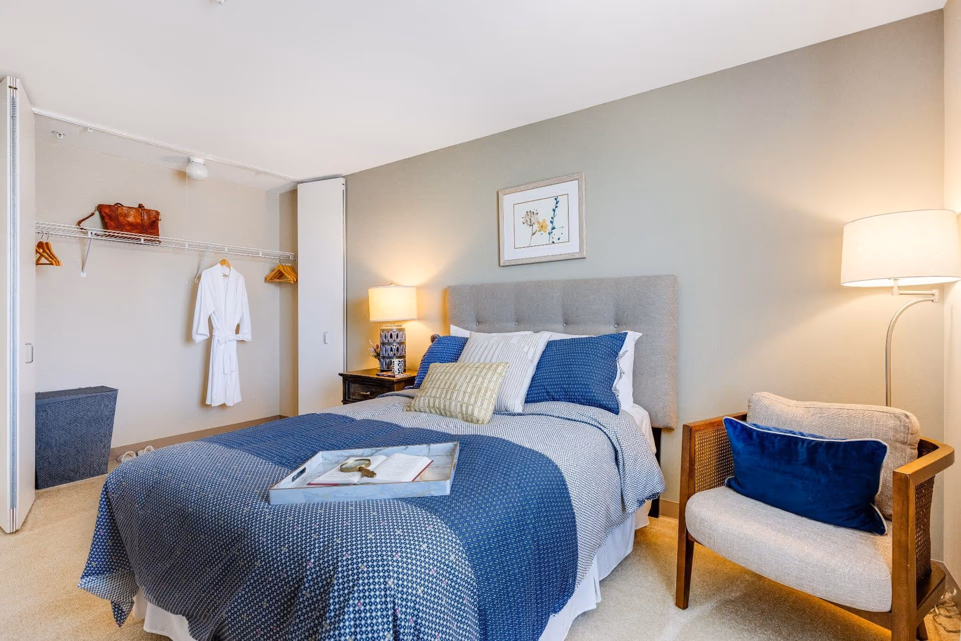 A cozy bedroom with a neatly made bed featuring blue and white bedding and multiple pillows. A tray with a book and a magnifying glass rests on the bed. To the right of the bed is a cushioned armchair with a blue pillow and a floor lamp. On the left side, there is an open closet with a white robe hanging and a brown bag on the shelf. A framed floral picture hangs on the wall above the bed.