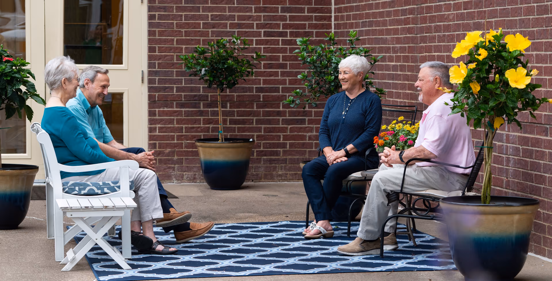 Four senior adults sitting and chatting on an outdoor brick patio with potted plants and a patterned rug.