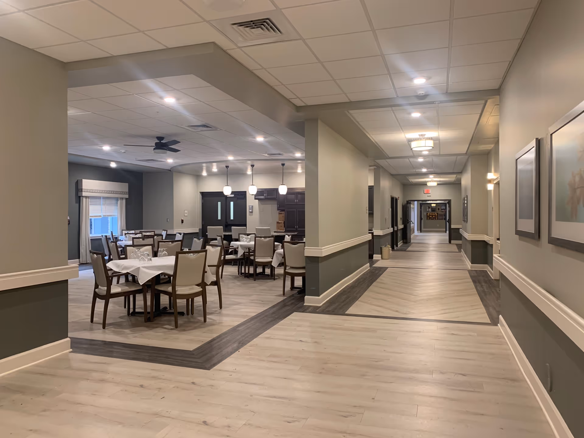 Interior view of a senior living facility showing a dining area with tables and chairs set with white tablecloths and glassware on the left, and a long hallway with framed artwork on the walls and ceiling lights on the right.