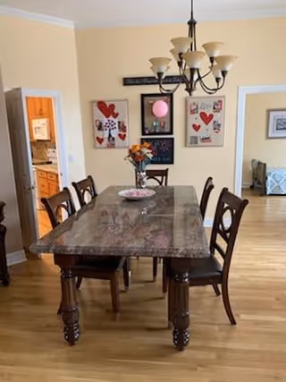 Dining room with a rectangular marble-top table surrounded by six wooden chairs with cushioned seats. A chandelier with multiple lights hangs above the table. On the wall behind the table, there are three framed artworks featuring hearts and other designs. A vase with flowers and a decorative plate are placed on the table. To the left, an open doorway leads to a kitchen area with wooden cabinets. To the right, another doorway leads to a room with a bed partially visible.