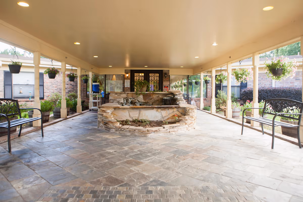 Covered outdoor patio area with stone flooring and a central stone water feature surrounded by benches and hanging flower pots. The area is well-lit with ceiling lights and has a garden visible in the background.