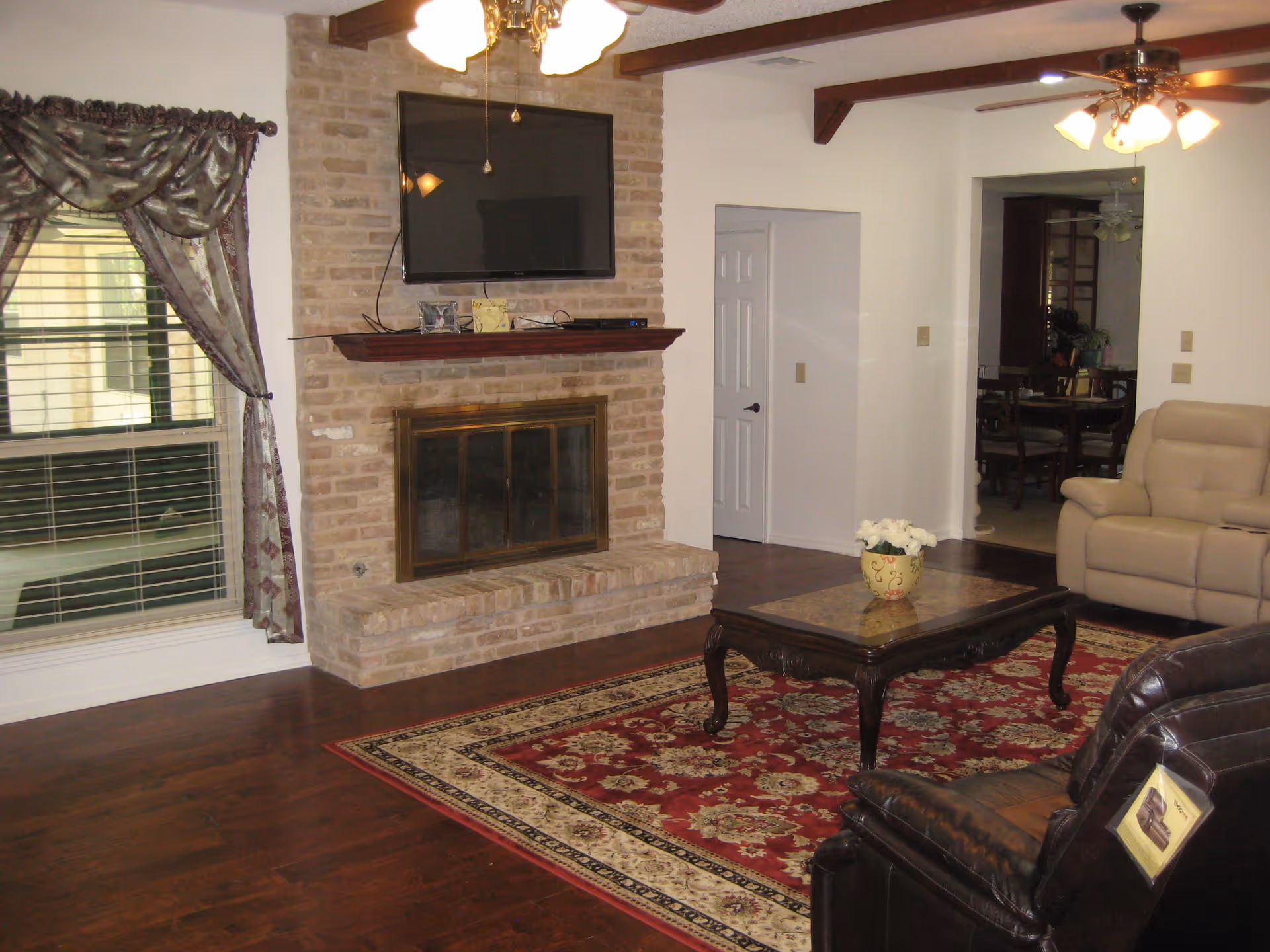 A cozy living room with a brick fireplace, a mounted flat-screen TV above the mantel, a large window with patterned curtains, a wooden coffee table with a flower pot, a red patterned area rug, and leather recliner chairs. The room has wooden floors and ceiling fans with lights.