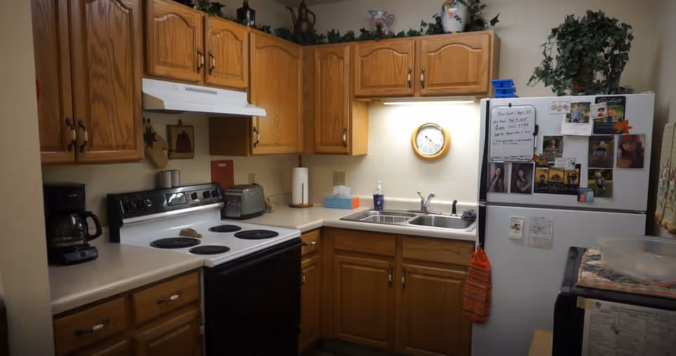 A compact kitchen with wooden cabinets, a white electric stove with four burners, a coffee maker, a toaster, a double sink, and a refrigerator covered with photos and notes. There is a clock on the wall above the sink and some greenery on top of the cabinets.