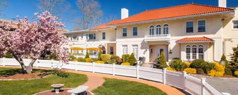 Two-story beige nursing home with a red tile roof, white fence, and blooming pink tree.