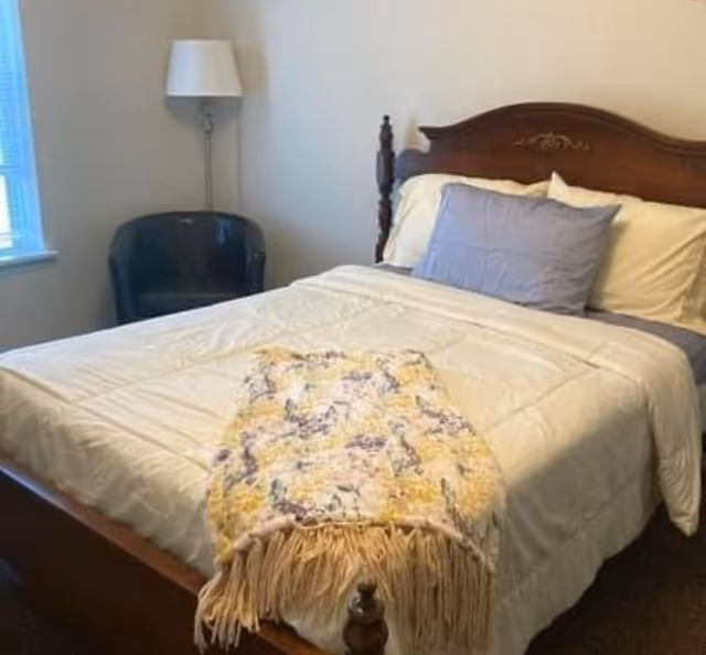 A bedroom with a wooden bed frame featuring a carved headboard. The bed is made with white bedding, a gray pillow, and a floral-patterned throw blanket with fringes at the foot. To the left of the bed is a black armchair and a floor lamp with a white lampshade. A window with blinds is partially visible on the left wall.