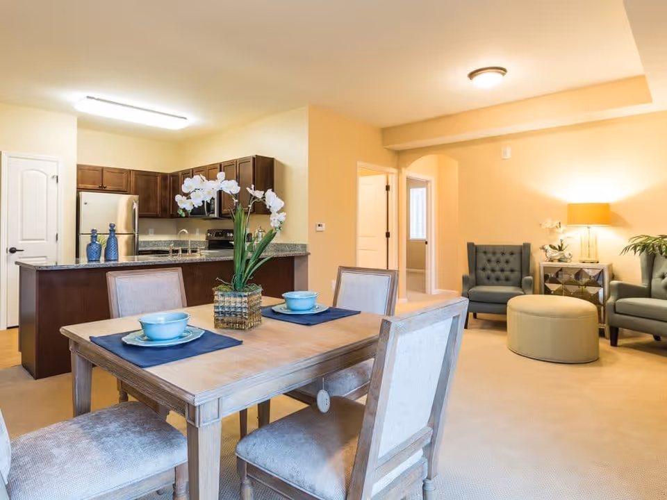 A bright and cozy open-concept living and dining area in a senior living facility. The foreground features a wooden dining table set with four upholstered chairs, two place settings with blue bowls and plates, and a decorative vase with white flowers. In the background, there is a kitchen with dark wood cabinets, granite countertops, and stainless steel appliances. To the right, a living area includes two blue armchairs, a round ottoman, a mirrored side table with a lamp, and a potted plant. The walls are painted a warm beige color, and the room is well-lit with ceiling lights.