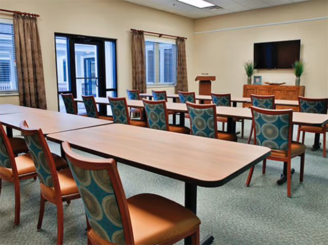 Bright indoor dining room with long tables, teal chairs, a podium, and wall-mounted TV.