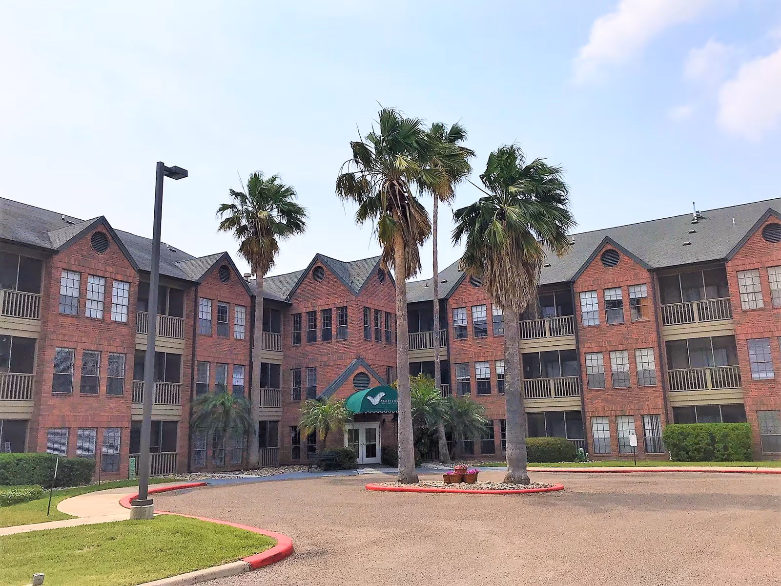 Exterior view of a three-story brick building with multiple windows and balconies, surrounded by palm trees and a circular driveway with a green awning entrance labeled Valley View Independent Living.