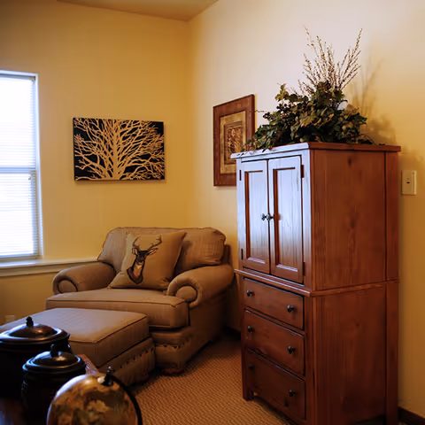 Cozy corner of a living room with a cushioned armchair and matching ottoman, a pillow with a deer design, a wooden cabinet with drawers, decorative plants on top, and framed artwork on the walls. A window with blinds lets in natural light.