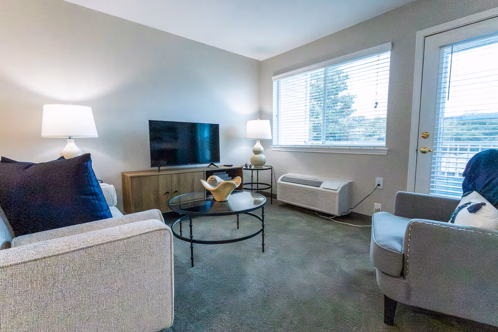 A bright furnished living room with a sofa and armchair, round glass coffee table, TV on a console, and a large window with blinds.