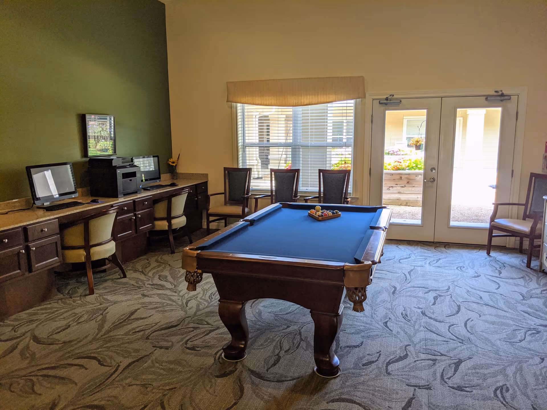 A recreational room with a blue felt pool table in the center, a row of computer workstations with chairs along the left wall, and several chairs near a window and glass double doors that lead outside. The room has a patterned carpet and a green accent wall.