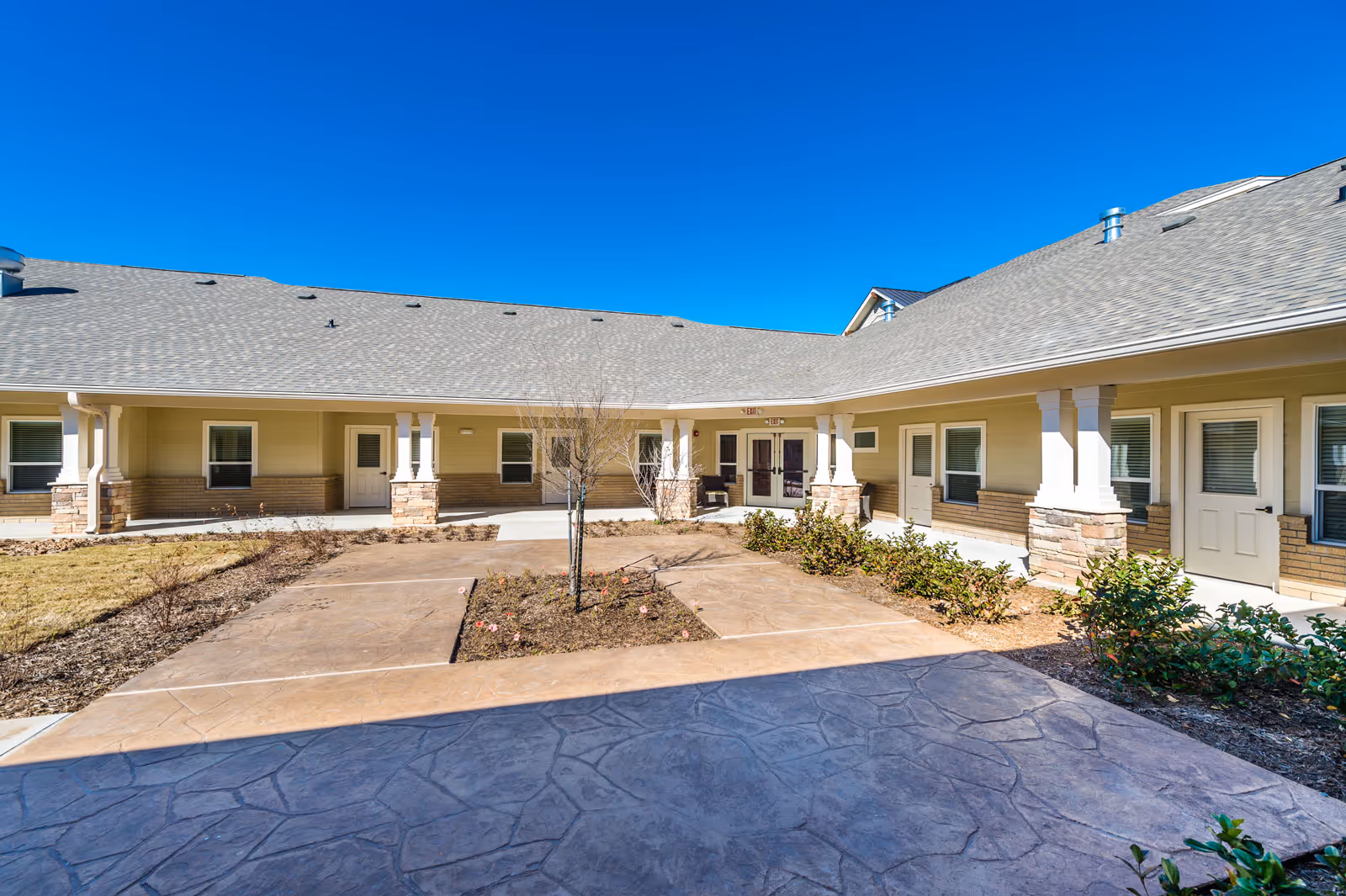 Outdoor courtyard area of a senior living facility with a paved walkway, small garden beds, and a single small tree in the center. The building surrounding the courtyard has beige siding, stone accents on the columns, multiple windows, and white doors under a gray shingled roof. The sky is clear and blue.