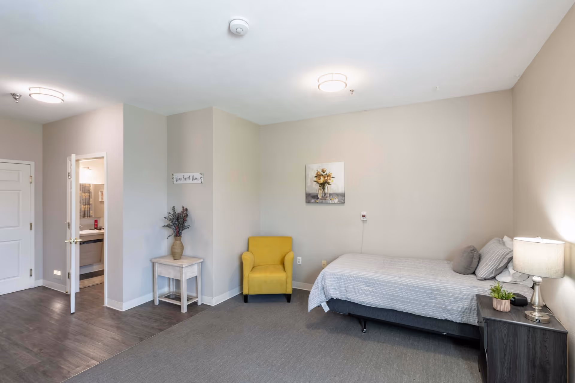 A simple senior bedroom with a single bed, bedside table and lamp, a yellow armchair, and an open bathroom door.