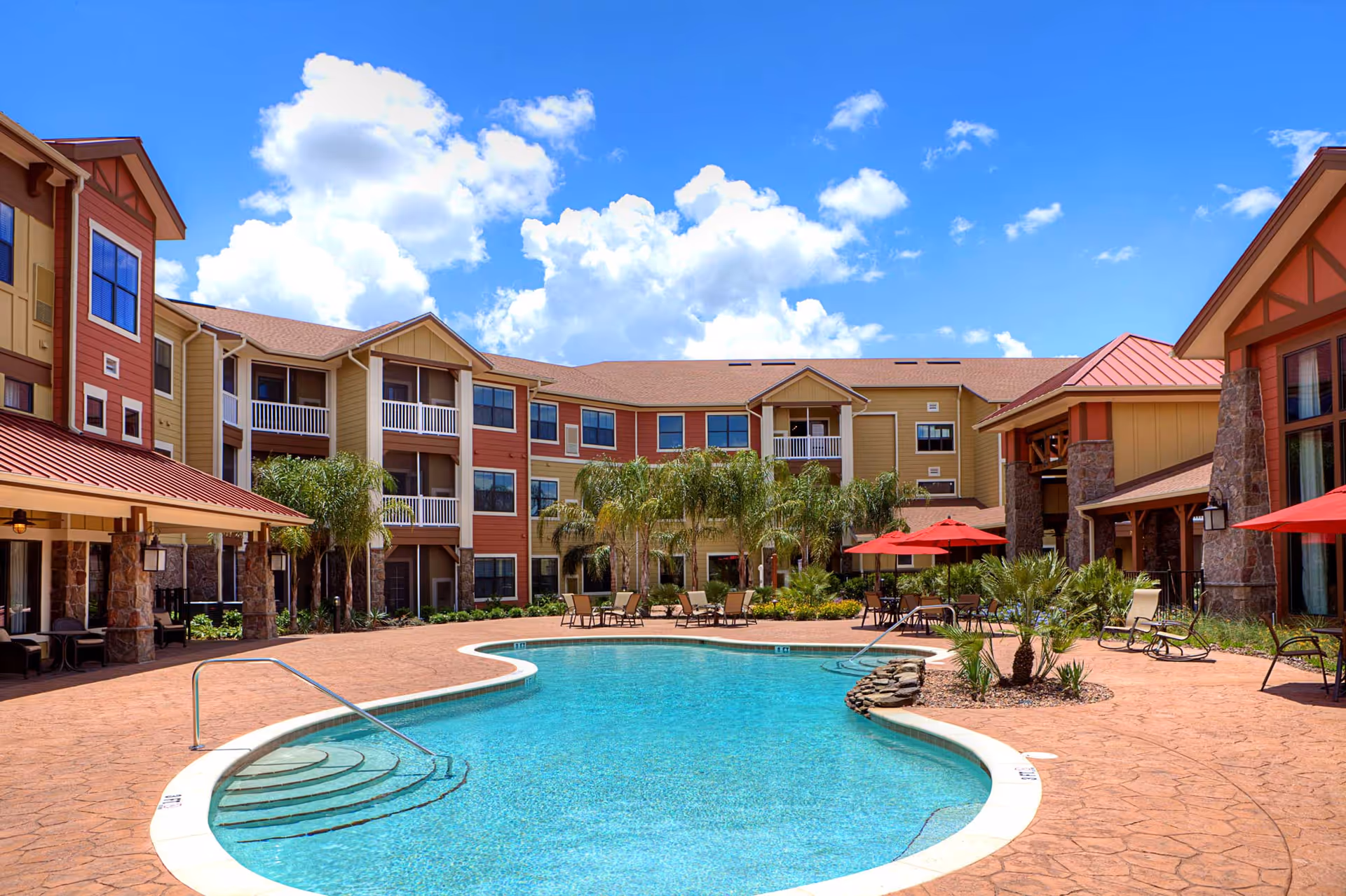 Outdoor swimming pool area at American House Wildwood with clear blue water, surrounded by a patio with lounge chairs, tables with red umbrellas, palm trees, and a multi-story building with balconies under a bright blue sky with scattered clouds.