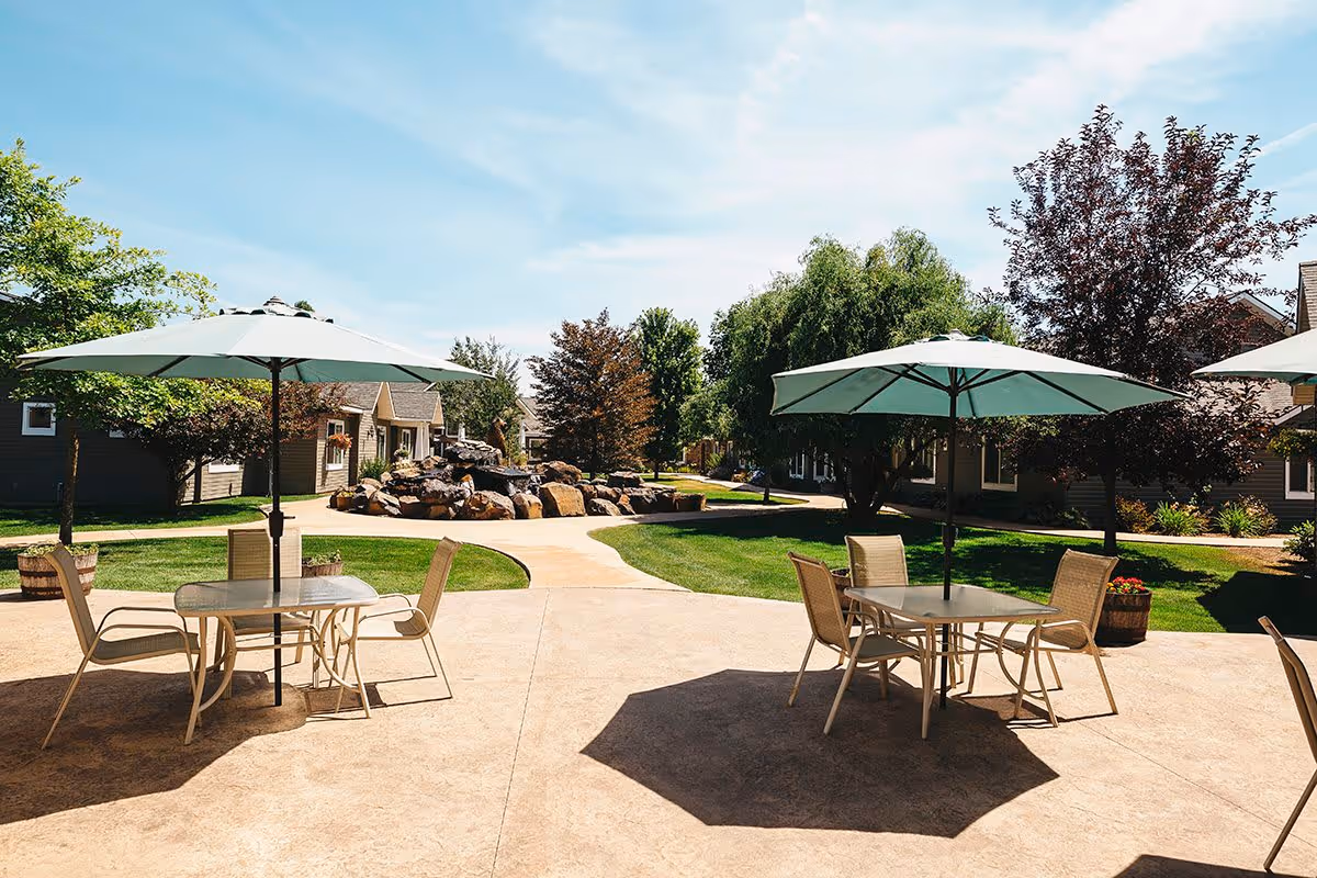 Outdoor patio area with tables and chairs under large umbrellas, surrounded by green grass, trees, and residential-style buildings under a clear blue sky.