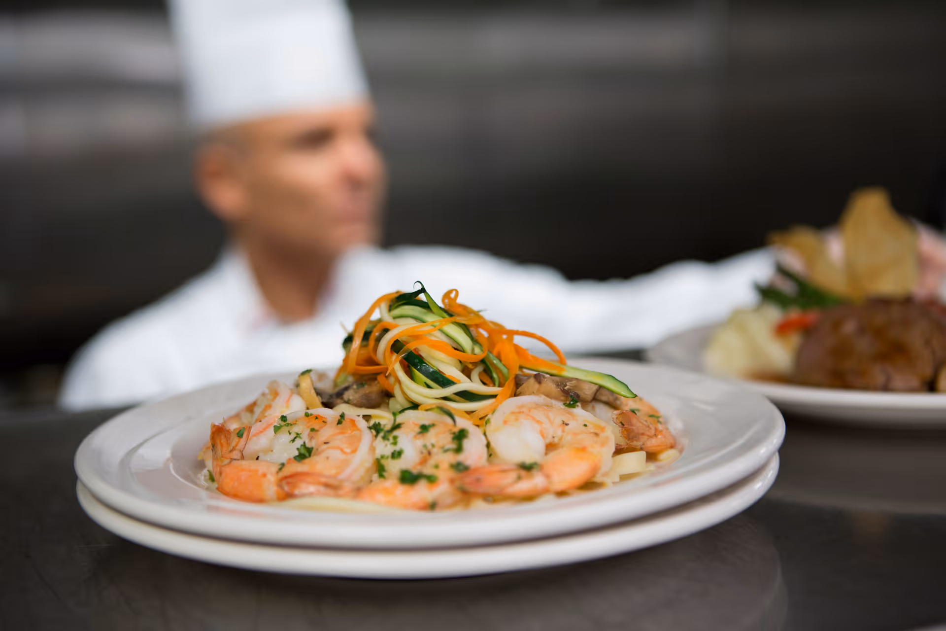 Close-up of a plate of cooked shrimp garnished with chopped herbs and topped with spiralized vegetables, with a blurred chef in a white uniform and hat in the background holding another plate of food.