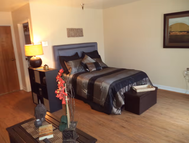 A bedroom with a neatly made bed featuring a gray and black striped comforter and multiple pillows. To the left of the bed is a black shelving unit with a lamp on top, and a wooden door is visible in the background. In the foreground, there is a coffee table with decorative items including a small statue, books, and a vase with red flowers. The room has wooden flooring and light-colored walls with a framed picture hanging on the right wall.