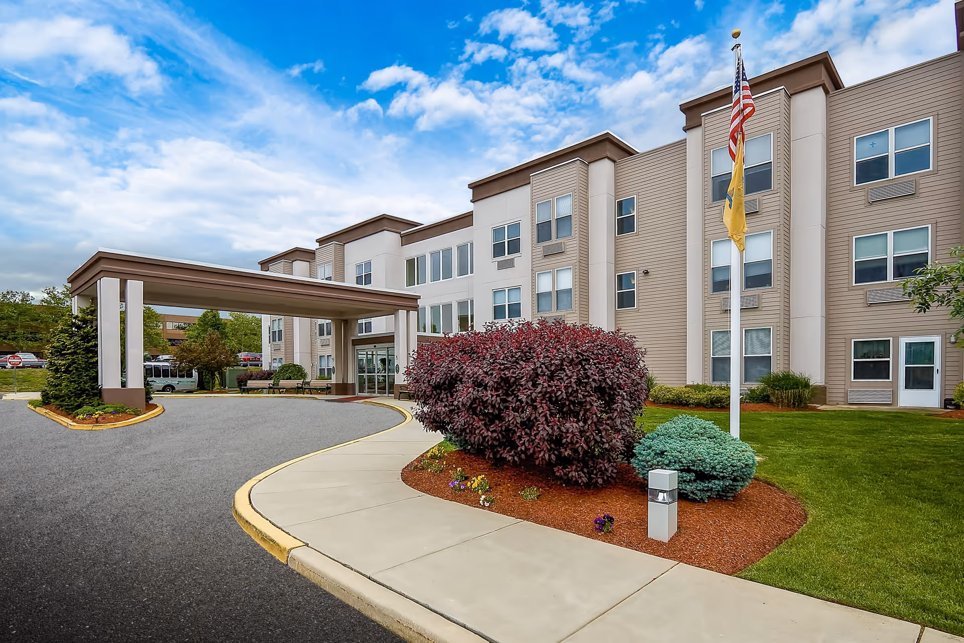 Exterior view of a three-story senior living facility building with beige siding and multiple windows. There is a covered entrance with a driveway and a sidewalk leading to the entrance. The landscaped area includes green grass, bushes, and a flagpole with an American flag and another flag beneath it. The sky is partly cloudy.
