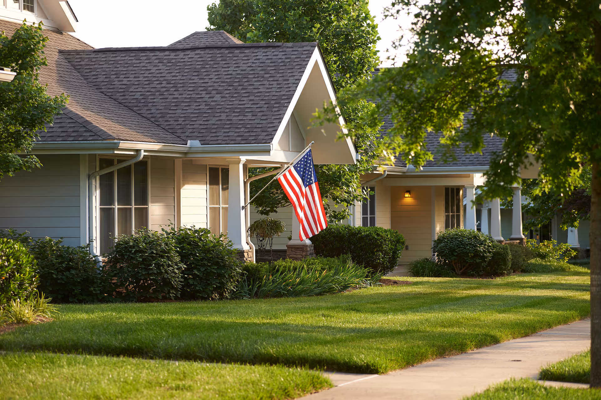 View of a residential-style building with a well-maintained lawn and bushes in front. An American flag is displayed on a porch with white columns. Trees provide shade along a sidewalk in a peaceful neighborhood setting.