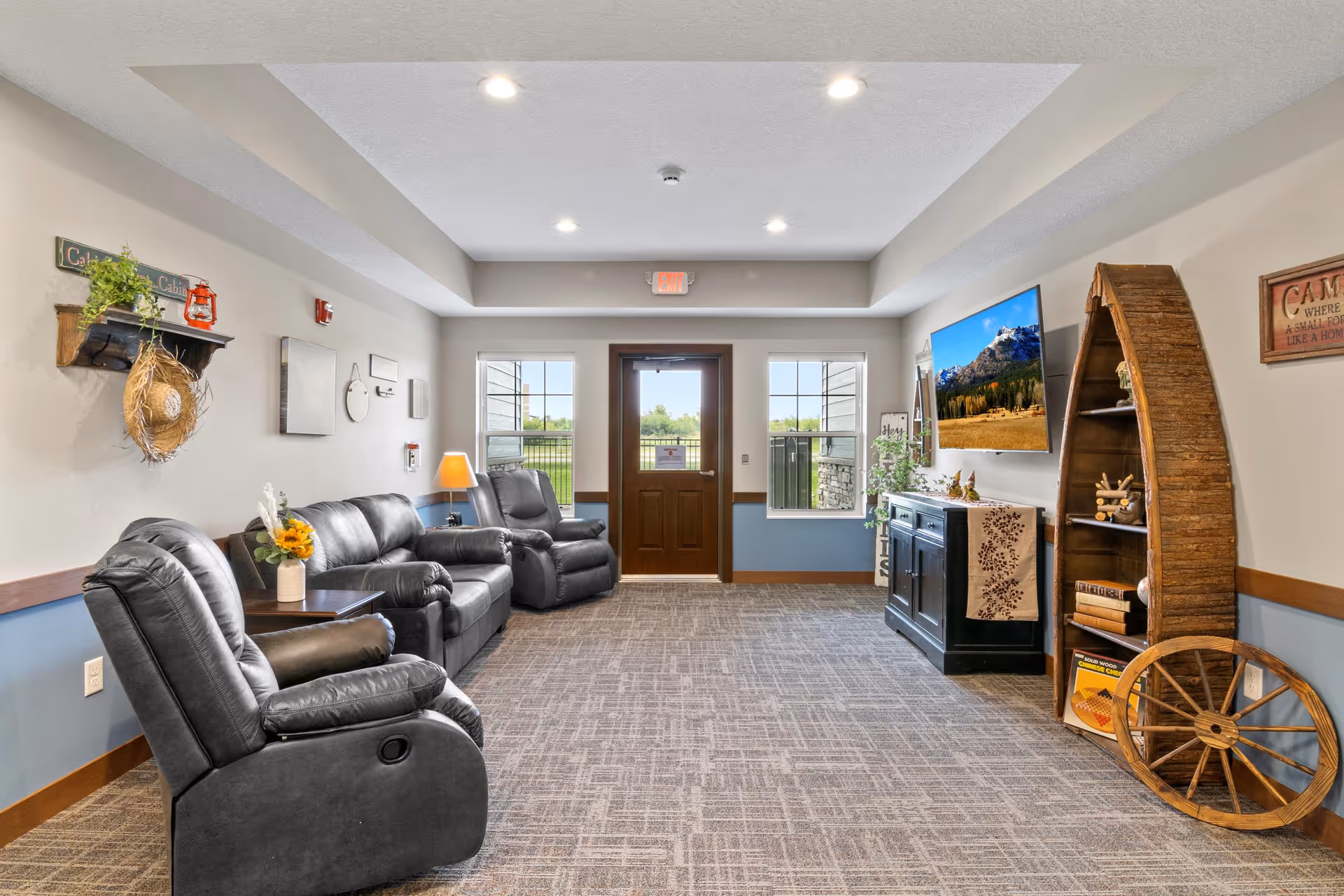 A cozy living room area with three black leather recliners arranged along the left wall. The room has a carpeted floor, beige walls with blue wainscoting, and a wooden door with two windows on either side letting in natural light. On the right wall, there is a flat-screen TV mounted above a black cabinet, next to a wooden boat-shaped bookshelf filled with decorative items and books. The room is decorated with a small shelf holding a plant and a hat, a lamp, and wall art, creating a warm and inviting atmosphere.