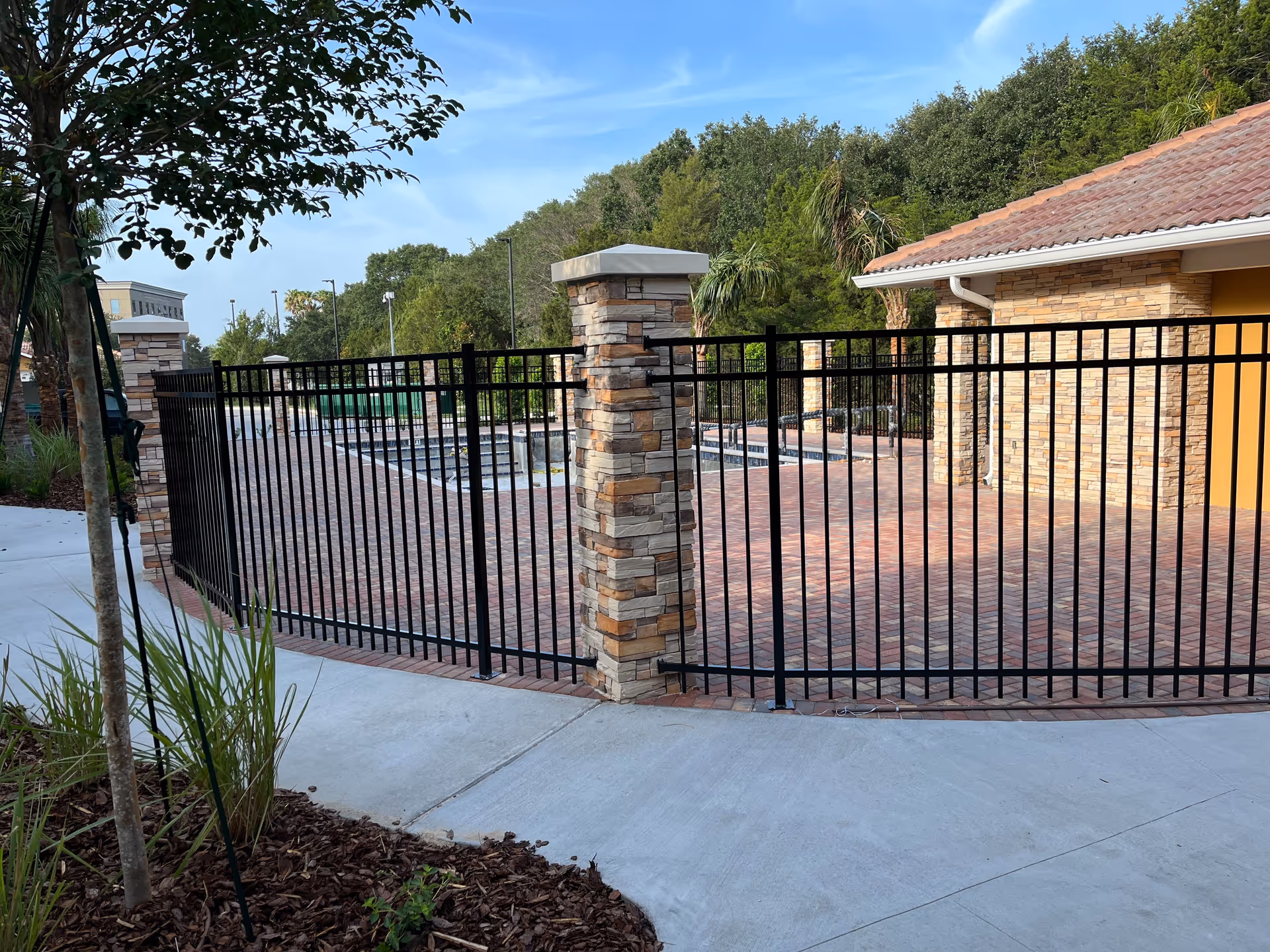 Black metal fence with stone pillars enclosing a paved courtyard and pool area with trees and buildings beyond.