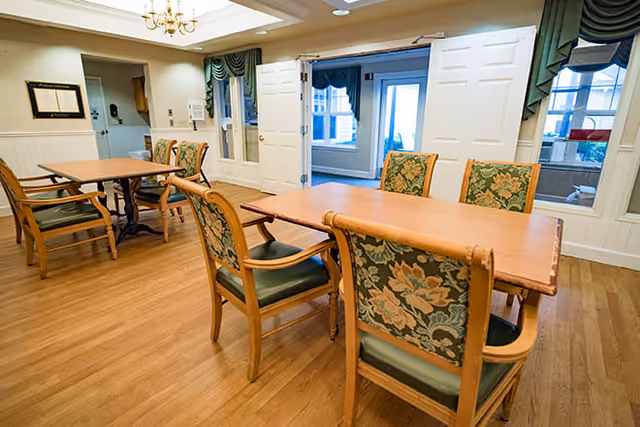 A bright dining area with wooden floors, two wooden tables, and several chairs with floral patterned upholstery. The room has white walls with wainscoting, a chandelier, and large windows with green valances. Double doors open to another room with more windows.