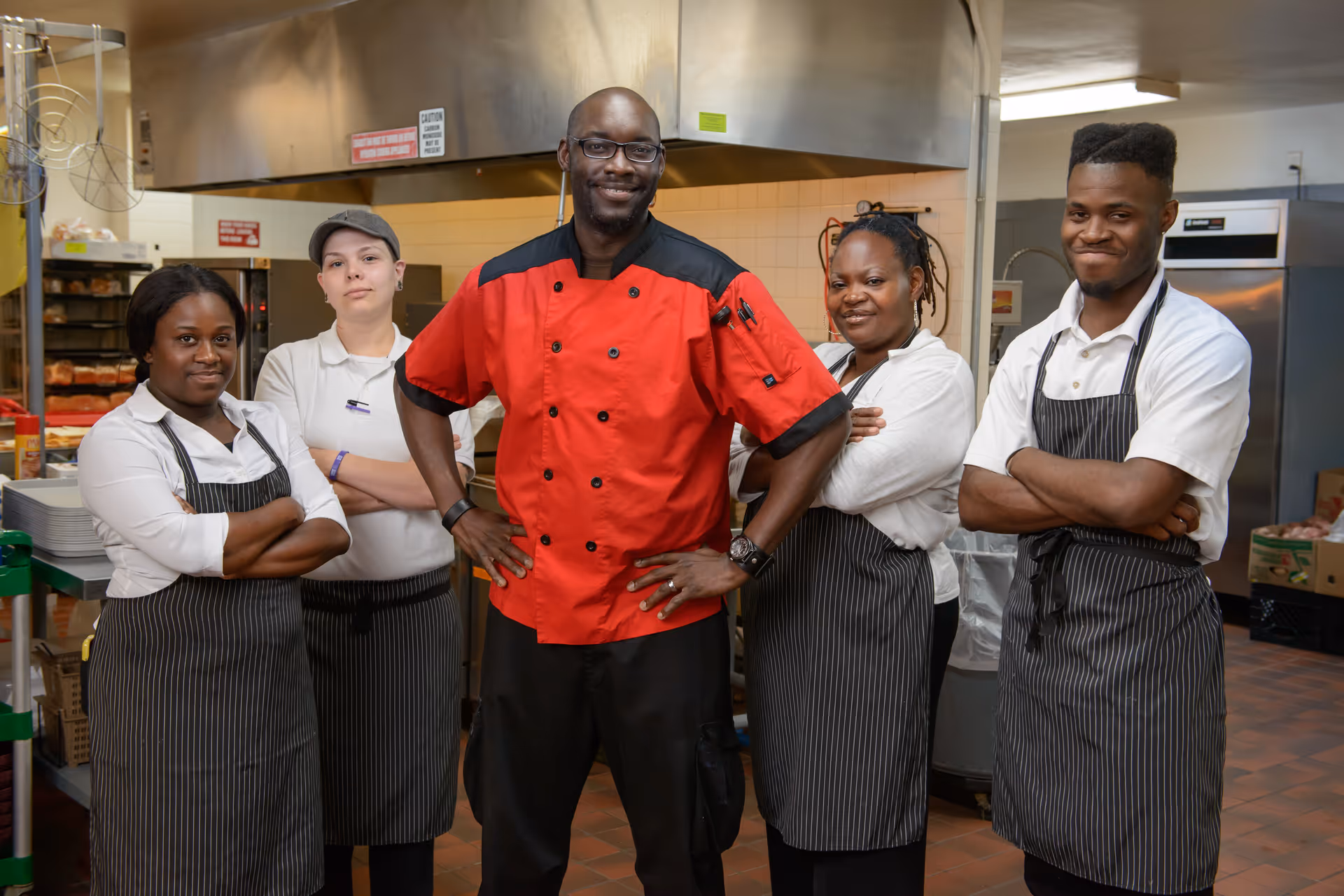 A group of five kitchen staff members standing together in a commercial kitchen. The central figure is a man wearing a red chef's jacket and glasses, with his hands on his hips. The other four individuals, two men and two women, are wearing white shirts and black striped aprons, standing with their arms crossed and smiling.