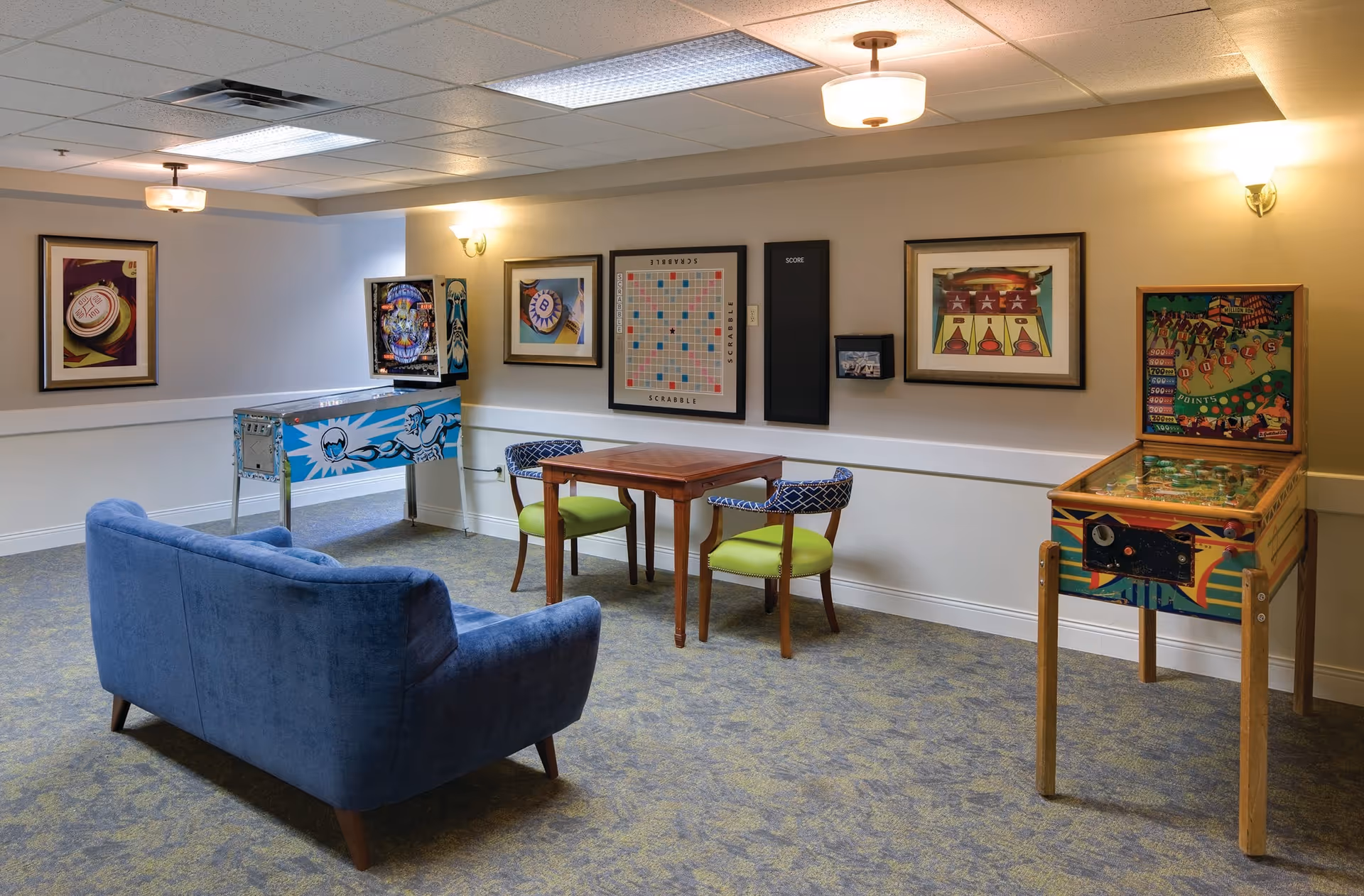 A cozy game room with a blue couch facing a wooden table with two green and blue patterned chairs. On the walls are framed pictures, including a large Scrabble board and a score board. The room also features two vintage pinball machines, one on each side of the table. The carpet is patterned in shades of blue and green, and the ceiling has recessed lighting and hanging light fixtures.