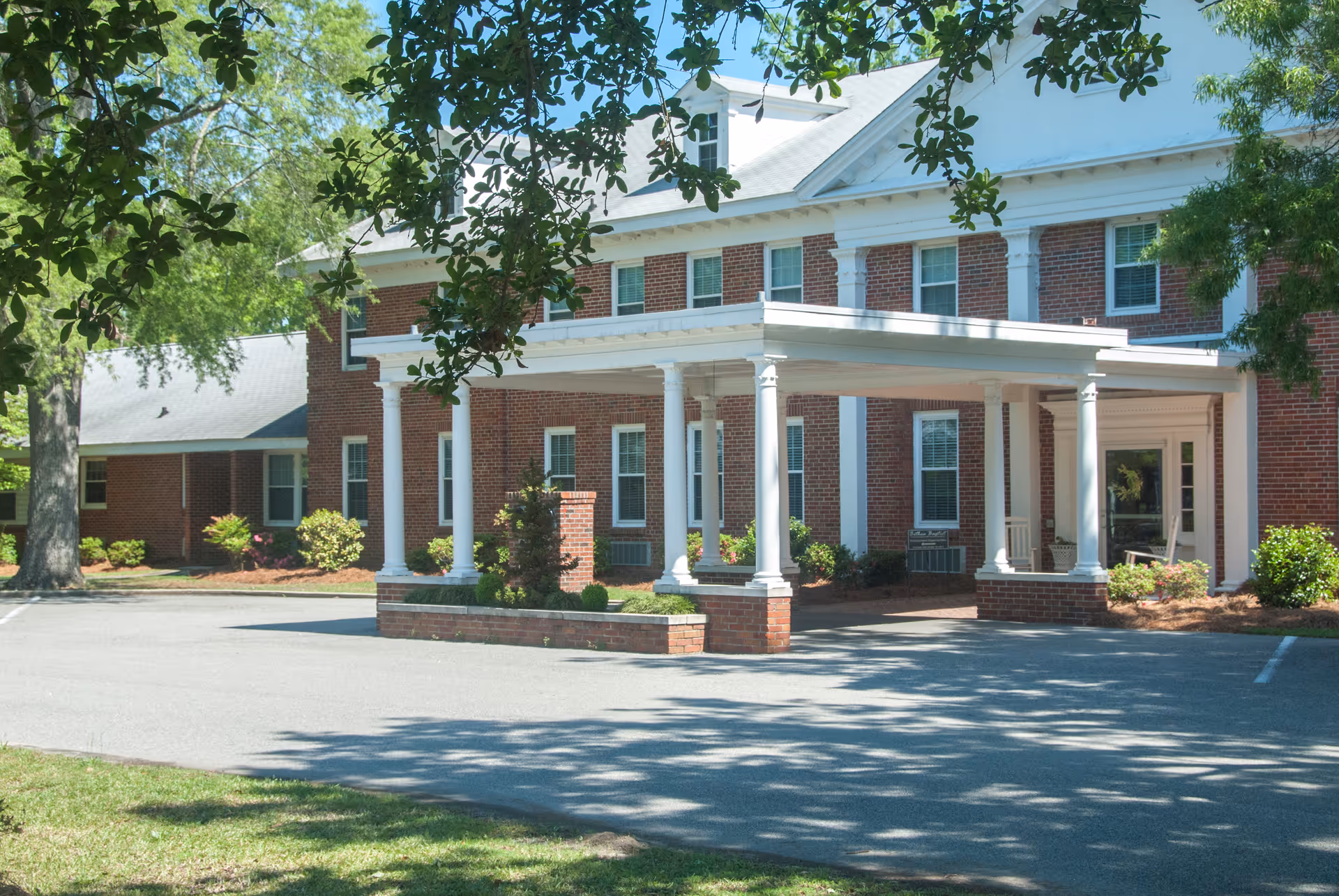 Exterior view of a two-story brick building with white columns and a covered entrance, surrounded by trees and greenery on a sunny day.