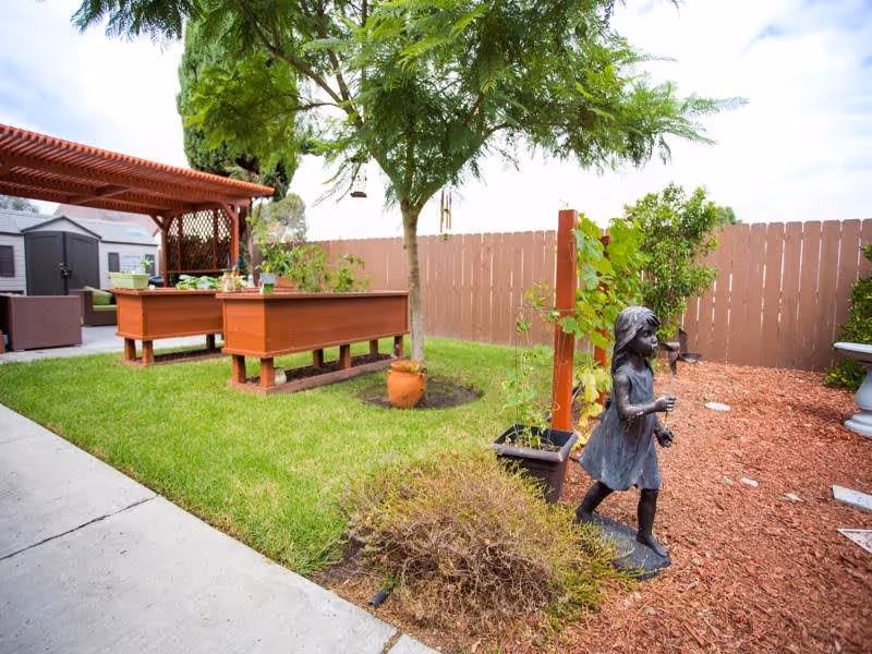 Outdoor garden area with green grass, a tree, raised wooden planter boxes, a small statue of a girl, and a wooden fence in the background. There is also a shaded seating area with outdoor furniture and a shed visible in the background.