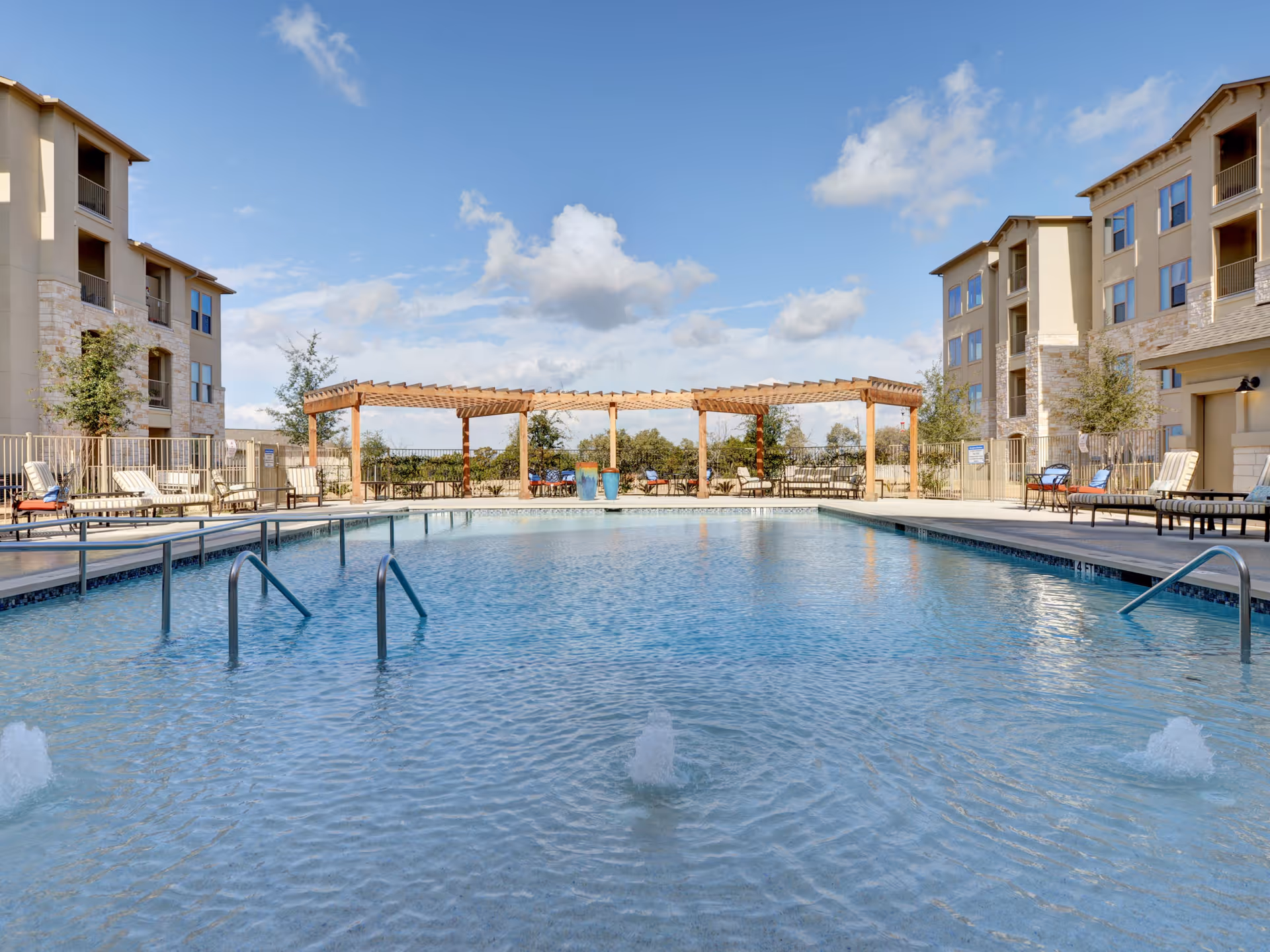 Outdoor swimming pool with clear blue water, surrounded by lounge chairs and tables under a wooden pergola. The pool area is enclosed by a fence, and there are multi-story residential buildings on either side under a partly cloudy sky.