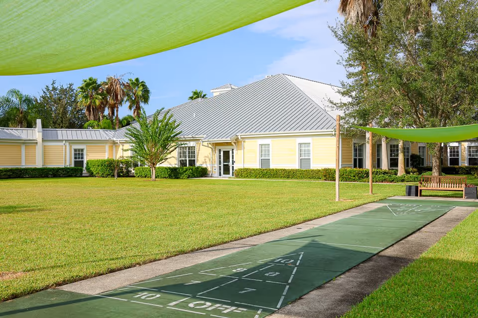 Outdoor shuffleboard court with a green playing surface and markings, situated on a lawn in front of a yellow building with a gray metal roof. There are palm trees and other greenery around, with green shade sails overhead and a wooden bench near the court.