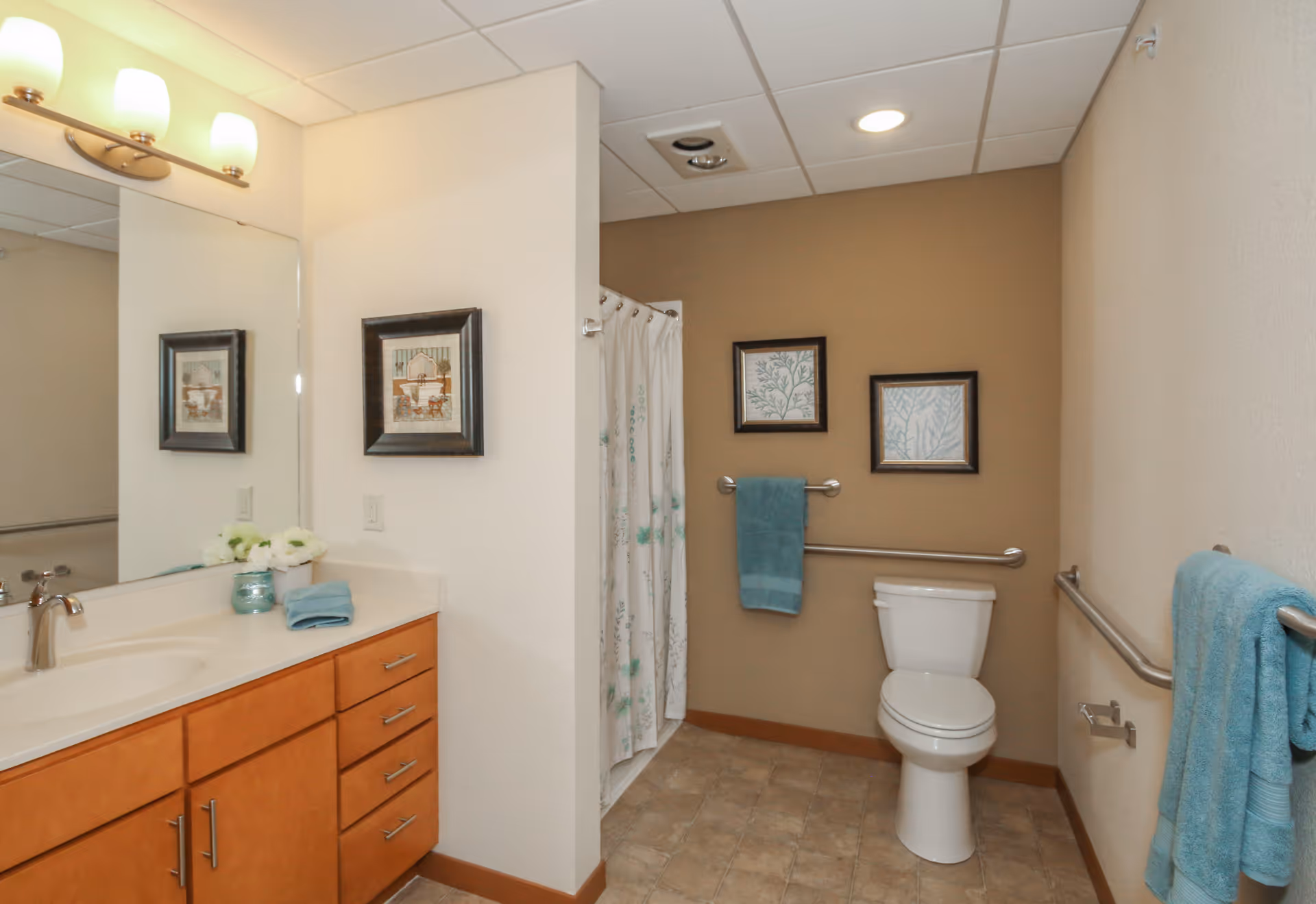 A clean and accessible bathroom with a white toilet, a shower with a curtain, a wooden vanity with a sink, a large mirror, and light fixtures above the mirror. The walls are painted beige and white, with two framed pictures and blue towels hanging on metal bars.