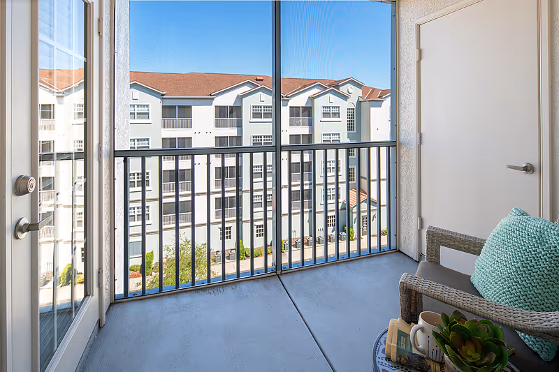 A small balcony with a wicker chair featuring a teal cushion, a small table with a plant, a cup, and a book. The balcony has a metal railing and overlooks a multi-story residential building with white and light blue exterior walls and a red roof under a clear blue sky.