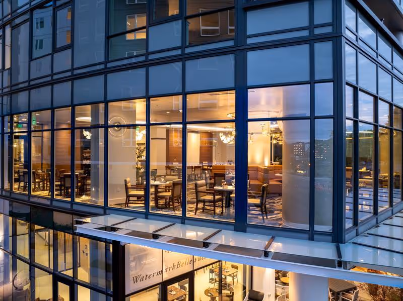 Exterior view of a modern building at dusk with large glass windows revealing a well-lit dining area inside, featuring multiple tables and chairs arranged neatly.