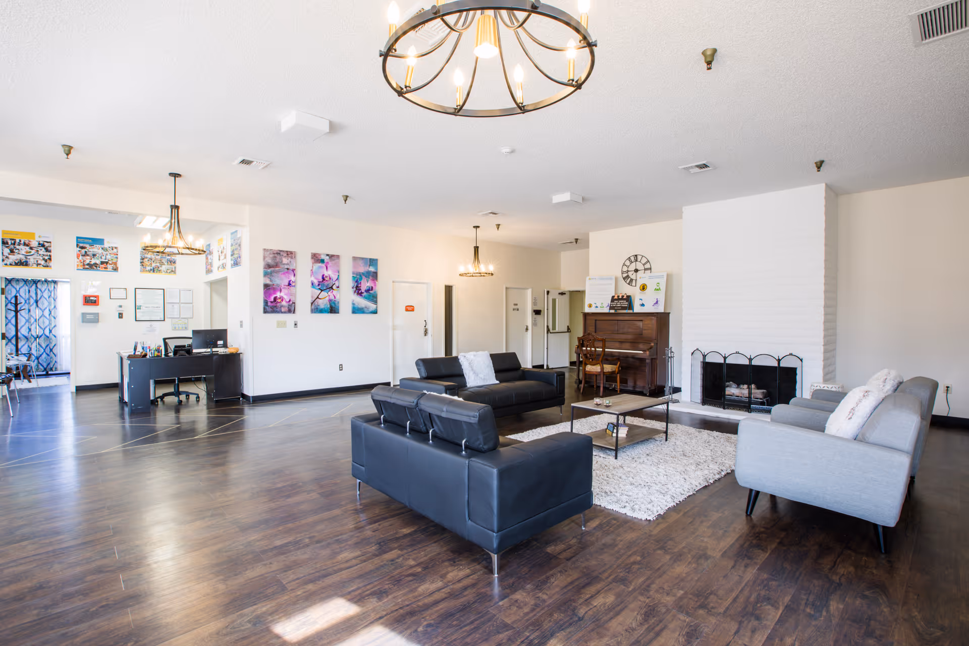 A spacious and well-lit living room area in a senior living facility with dark wood flooring, two black leather sofas, a gray fabric sofa, a white rug, a coffee table, a piano against the wall, and a white brick fireplace. The walls are decorated with colorful artwork and a large clock. There are two chandeliers hanging from the ceiling and a desk area visible in the background.