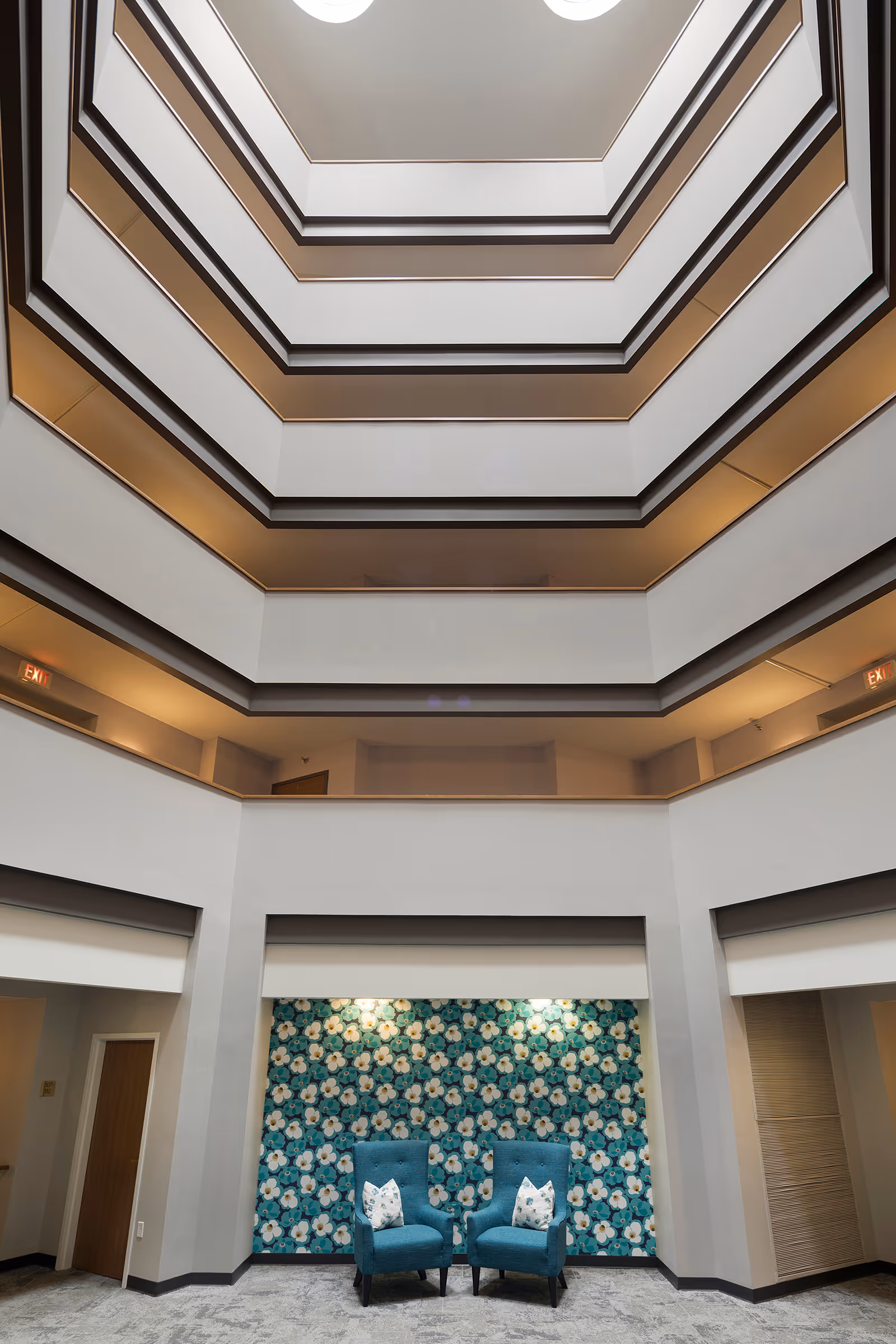 Interior view of a multi-story atrium with white walls and dark trim. At the bottom, there are two blue armchairs with floral patterned pillows placed in front of a wall covered with blue floral wallpaper. The ceiling has a skylight allowing natural light to enter.