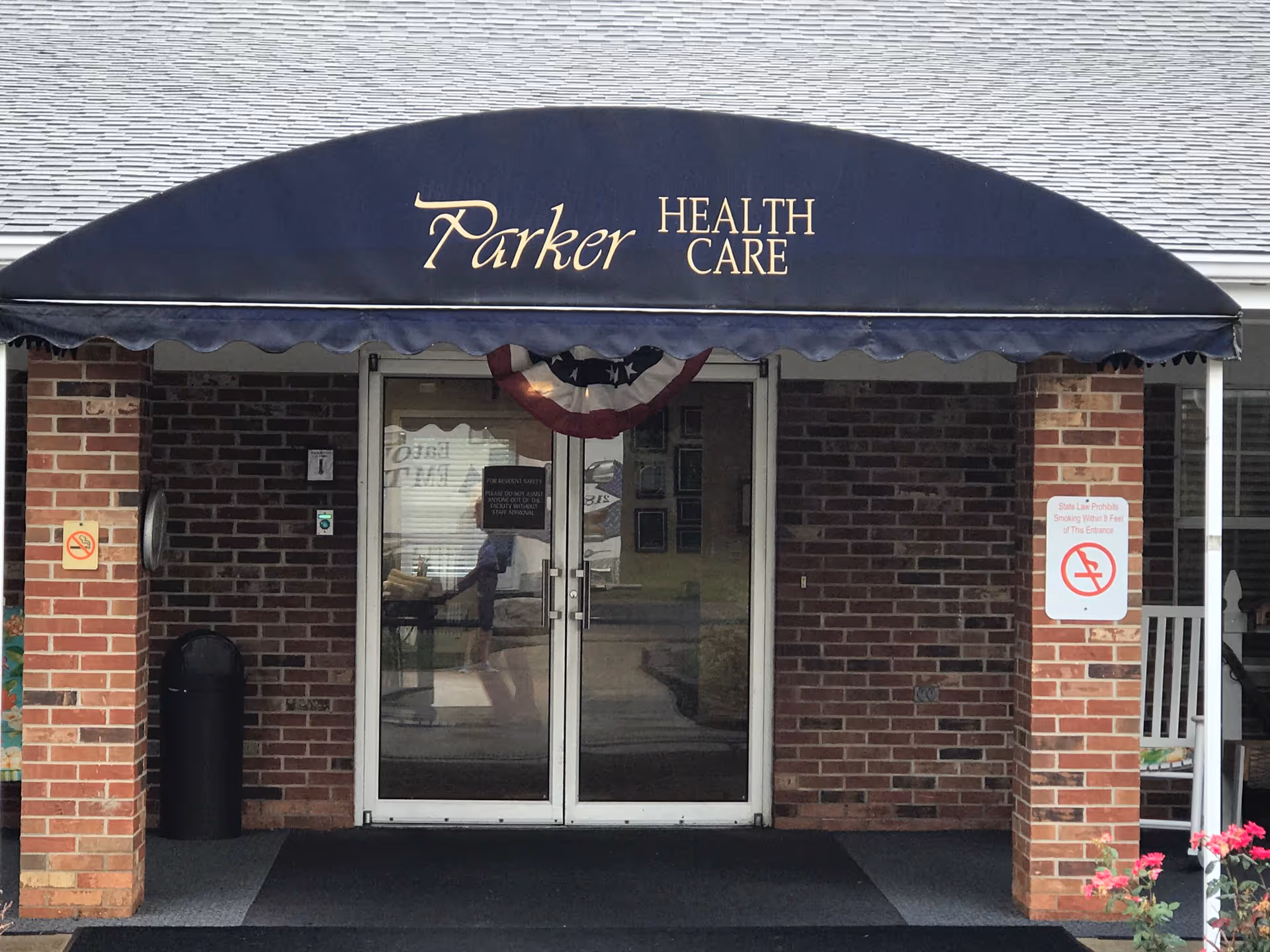Entrance to Parker Health Care facility with a blue awning displaying the name. The entrance has glass double doors framed by brick walls, with a no smoking sign on the right pillar and a trash can on the left side. There is a patriotic bunting decoration above the doors.