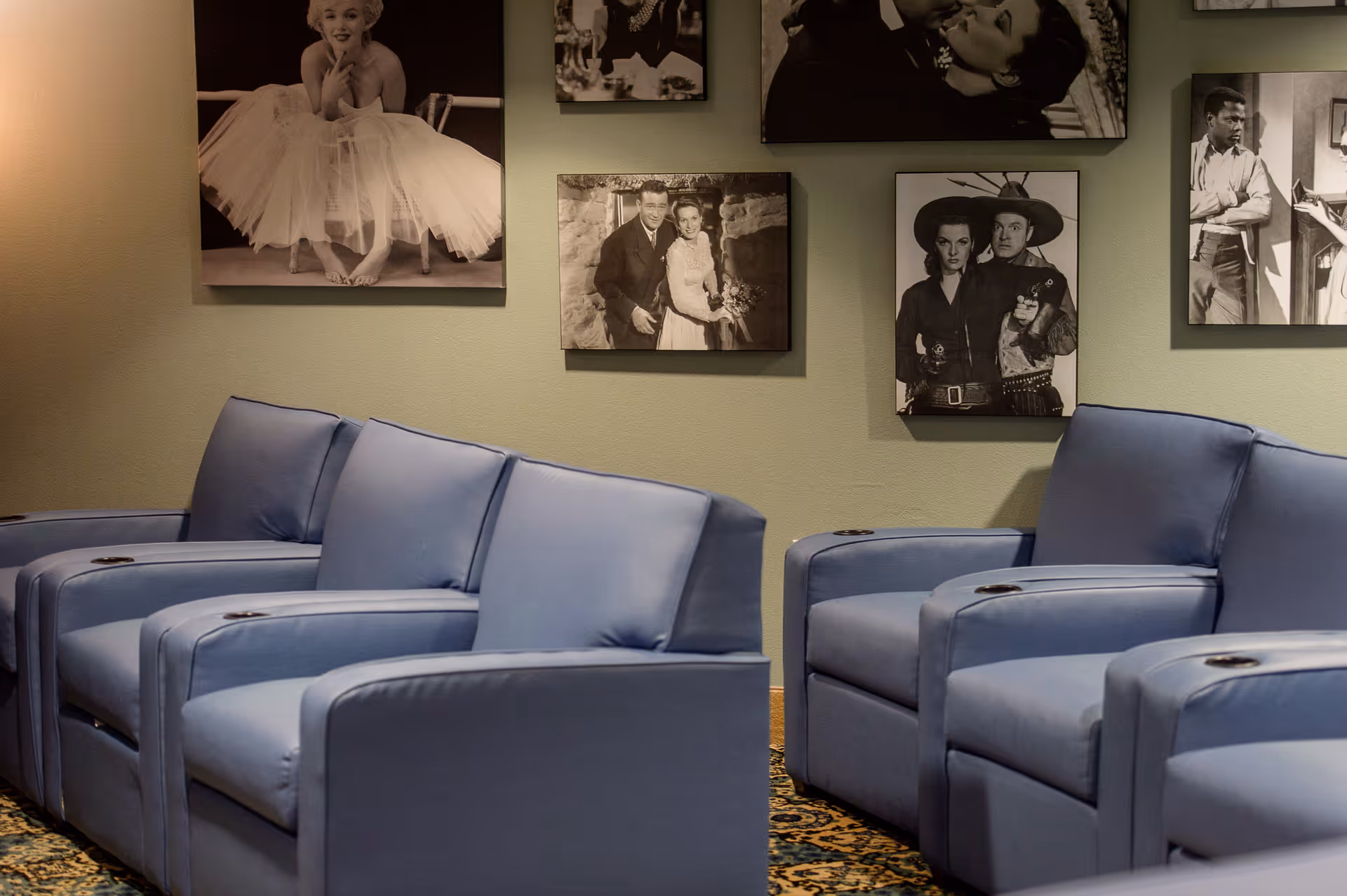 A row of blue cushioned armchairs with cup holders in a room decorated with black and white vintage movie posters on the wall.