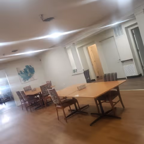 A spacious room with wooden flooring featuring two rectangular wooden tables and several chairs with patterned upholstery. The walls are light-colored with a piece of abstract artwork hanging on one side. There is a doorway leading to another room and a vent on the wall.